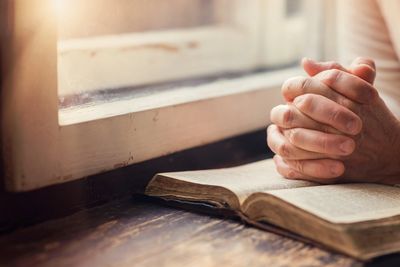 Hands clasped in prayer beside an open book near a window.