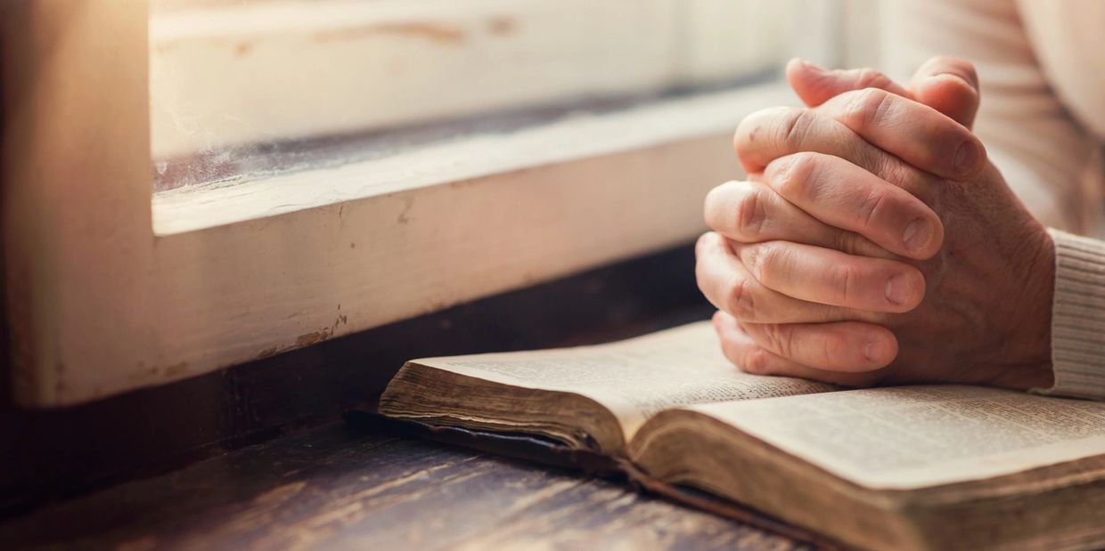 Folded hands on top of an open bible near a window.