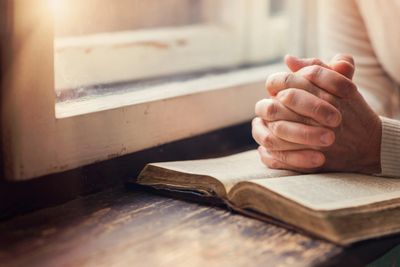 Praying hands on a Bible near a window. 