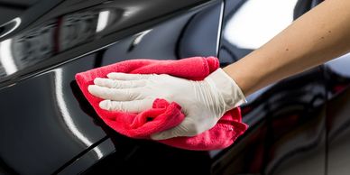 A gloved hand is polishing a car fender with a red terry cloth to remove the car wax. 