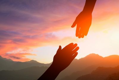 Silhouette of two hands, with one reaching up to the other with a sunset behind