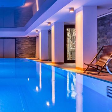 Indoor swimming pool with modern lighting and a wooden lounge chair.