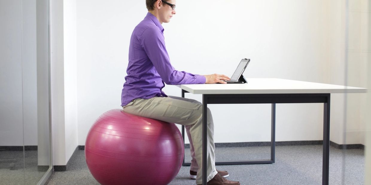 Man sitting on exercise ball working on tablet at desk.