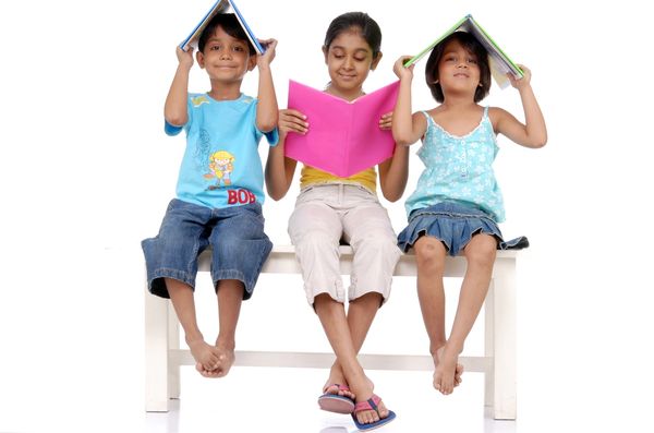 Three kids sitting on a bench, two holding books over their heads and one reading.