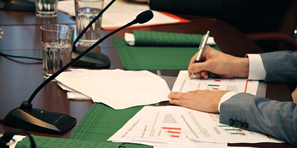 A person in a suit writing on documents at a conference table with microphones and glasses of water.