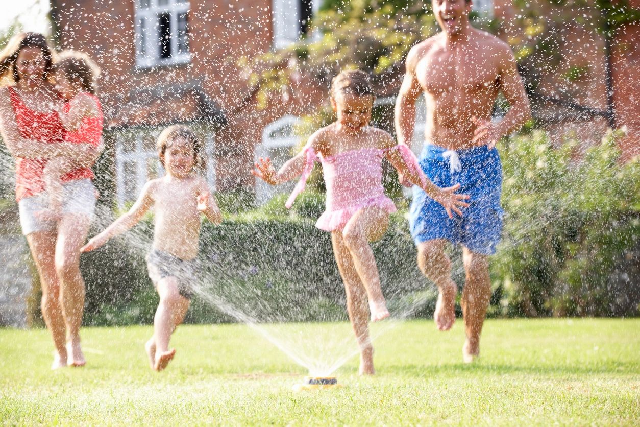 Family running around garden around sprinkler. 