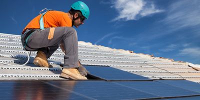 Worker installing solar panels on a roof under clear blue sky.