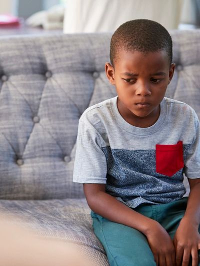 A sad young boy sitting alone on a couch, looking down.