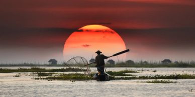 Fisherman silhouetted against a large setting sun on a calm water body.