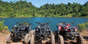 Three all-terrain vehicles parked by a lake with a forest in the background.