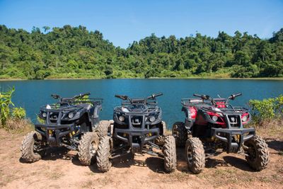 Three all-terrain vehicles parked by a lake with forested hills in the background.