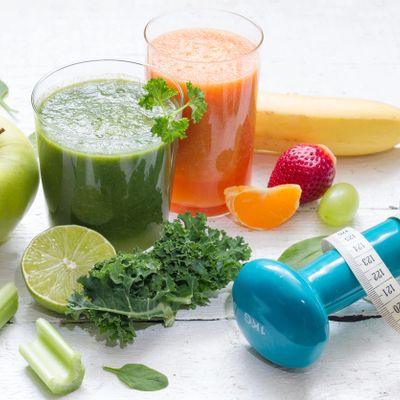 Two healthy smoothies surrounded by fresh fruits, vegetables, and a blue dumbbell with a measuring tape.