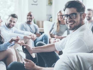Smiling man with glasses holding a tablet, surrounded by a diverse group of people in a casual setting.