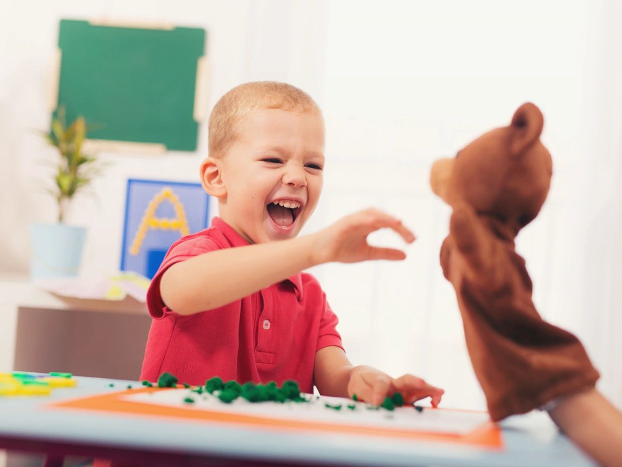 A joyful child playing with a bear hand puppet at a table.