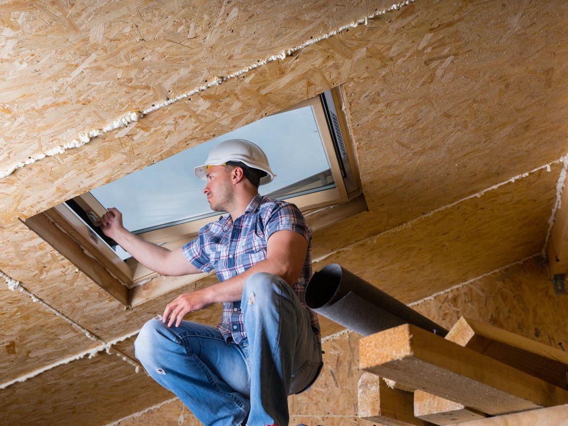 Man in a hard hat inspecting a skylight in an attic under construction.
