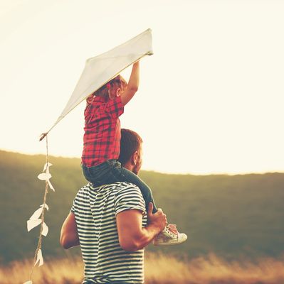 Child on father's shoulders holding a kite in a field at sunset.
