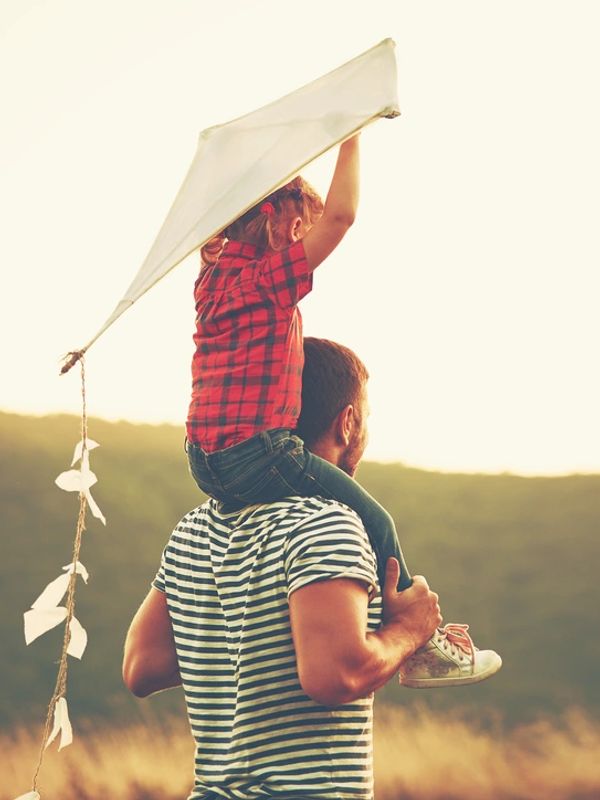 Child sitting on father's shoulders, holding a kite above her head.  They are going to fly the kite!