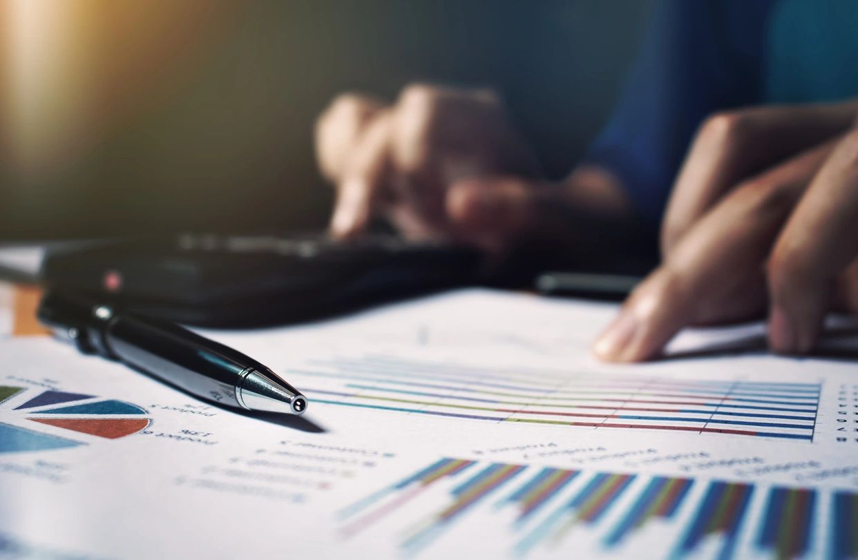 Pen on financial charts with person using calculator in background.