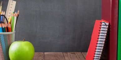 Green apple, colored pencils, and notebooks on a wooden desk with a blackboard background.
