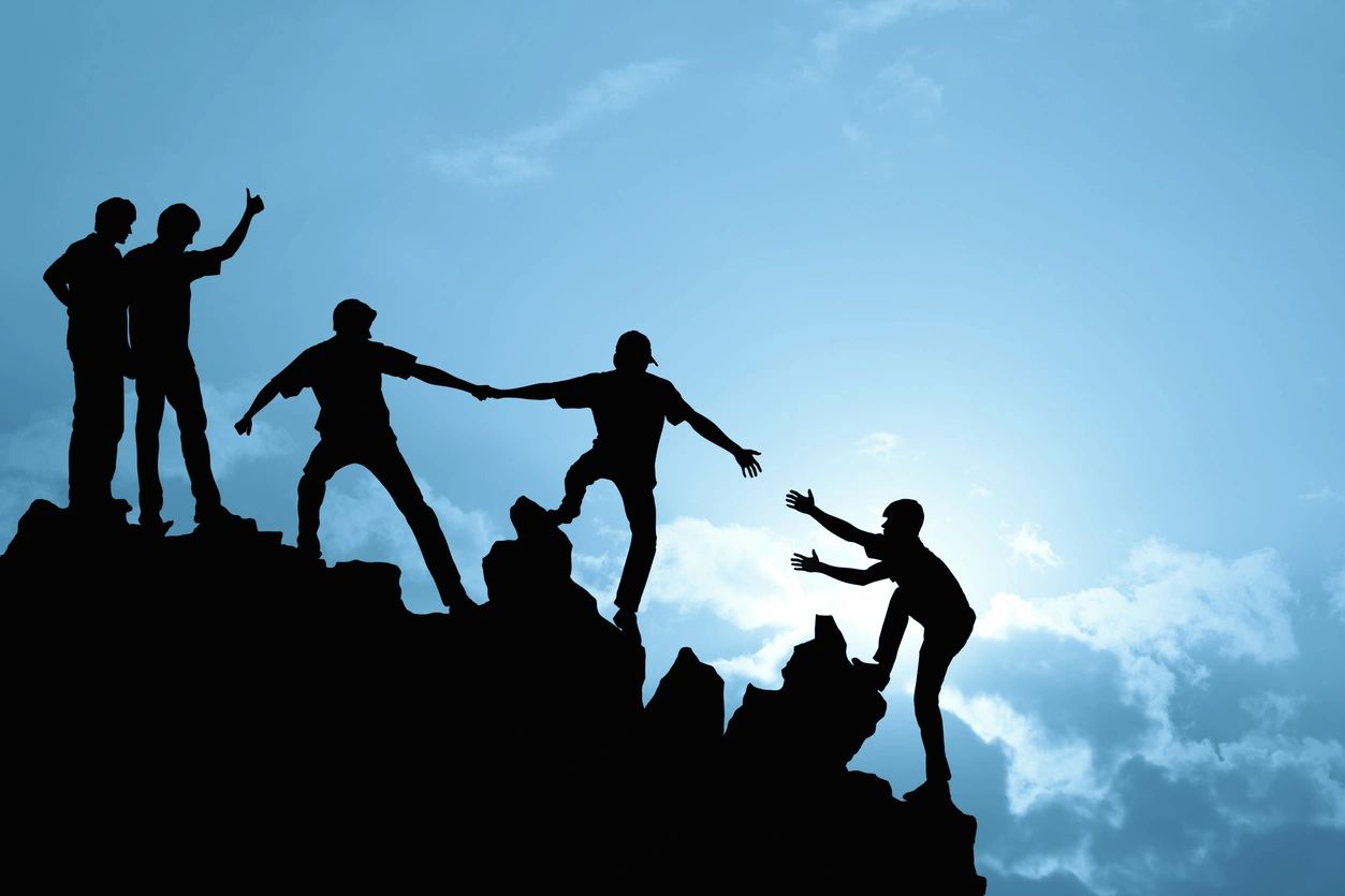 Silhouettes of people helping each other climb a rocky hill against a blue sky.