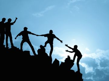 Silhouettes of people helping each other climb a hill at sunset.