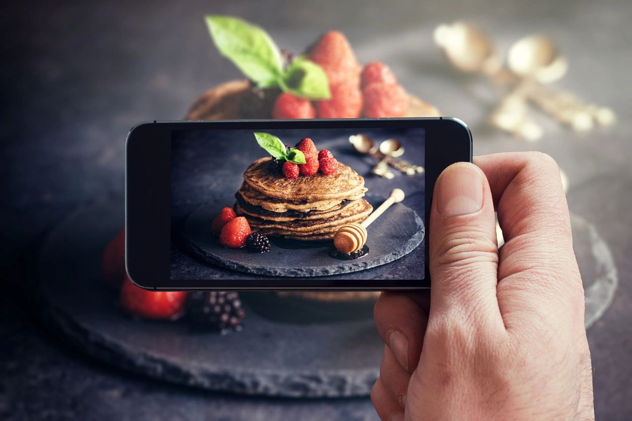 Hand holding smartphone capturing image of stacked pancakes topped with strawberries, raspberries, blackberries, and honey, emphasizing food photography in influencer marketing.