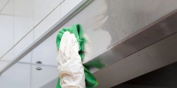 A gloved hand cleaning a stainless steel surface with a green cloth.