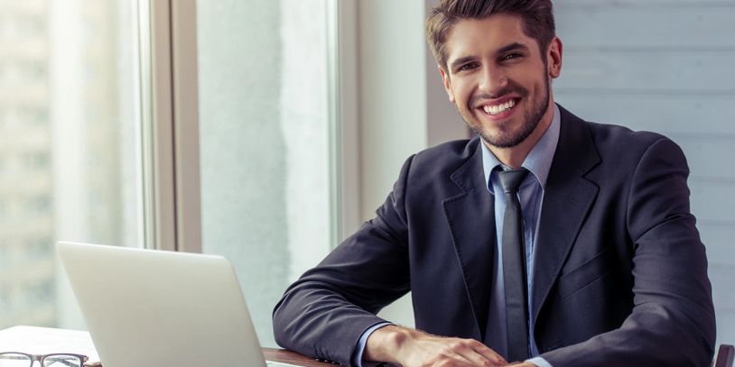 Smiling businessman in a suit working on a laptop at an office desk.
