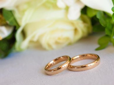 Two gold wedding rings placed on a white surface with white roses in the background.