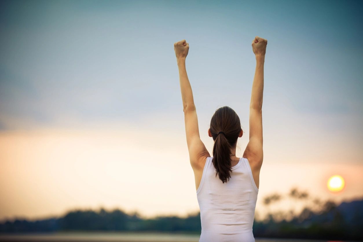 A young lady celebrates her win, fists in the air, overlooking the sun setting.