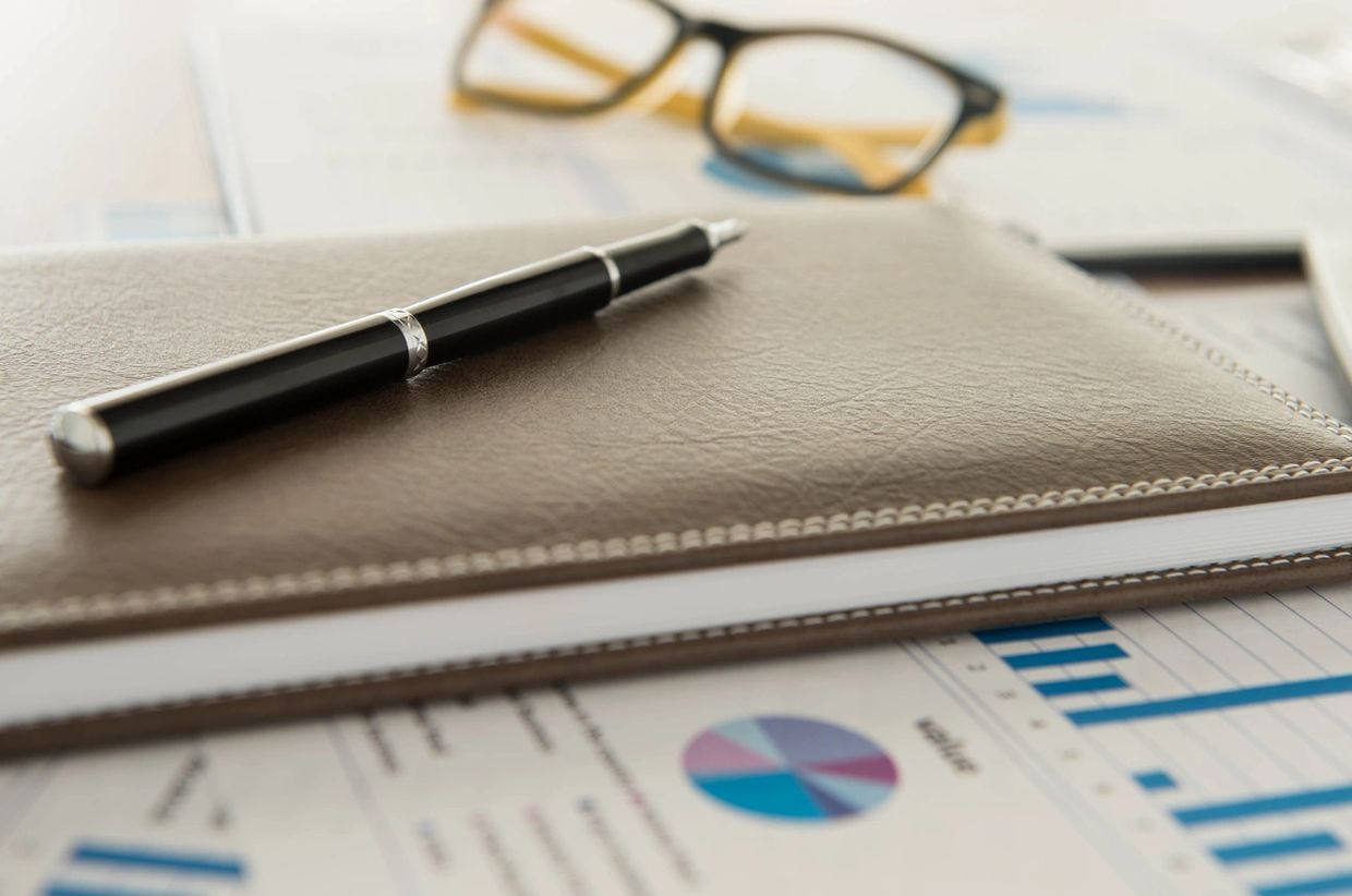 A pen resting on a leather notebook with glasses and charts in the background.