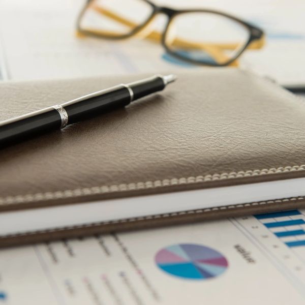 Pen on a leather notebook with glasses and financial charts in the background.