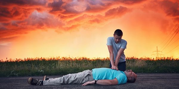 a man with a blue shirt laying outside on the floor and another is doing chest compressions.