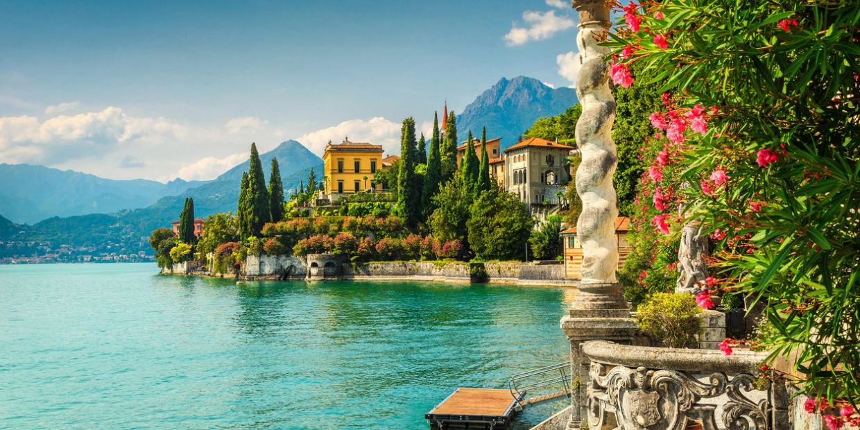 Scenic lakeside view with vibrant flowers, historic buildings, and mountains in the background.