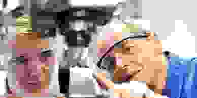 A workshop tutor pointing at a drill machine & showing it to a trainee both wearing safety glasses.