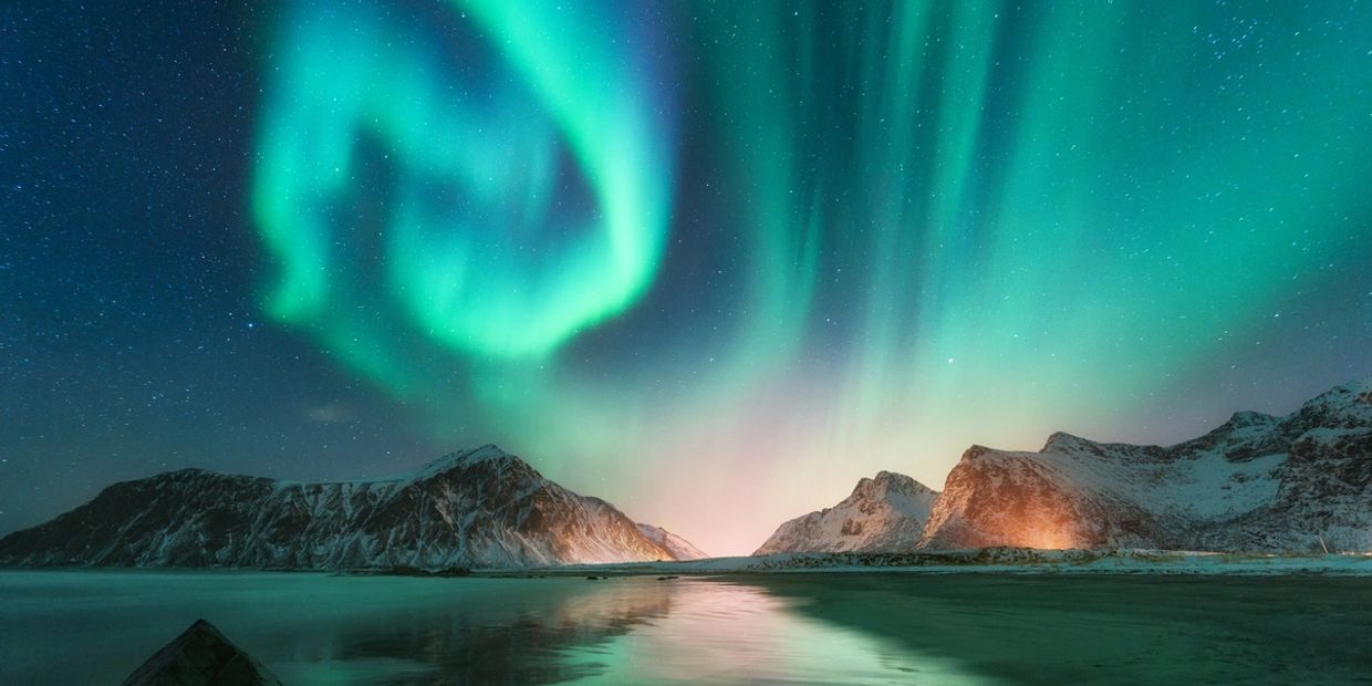 Northern lights swirl over snow-covered mountains and a reflective lake.