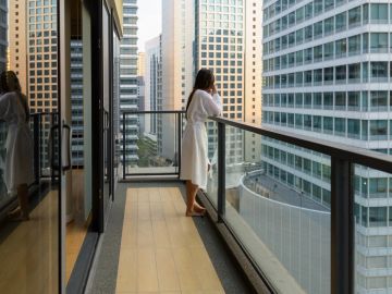 A woman in a white robe stands on a balcony overlooking city buildings.