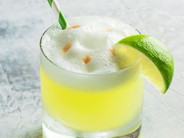 A green striped straw sits inside of an egg white whiskey sour, accompanied by a wedge of juicy lime