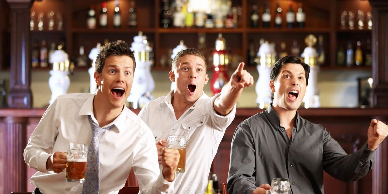 Three men excitedly cheer with beer mugs in a bar.