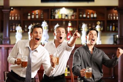 Three men excitedly watching something in a bar, holding beers.