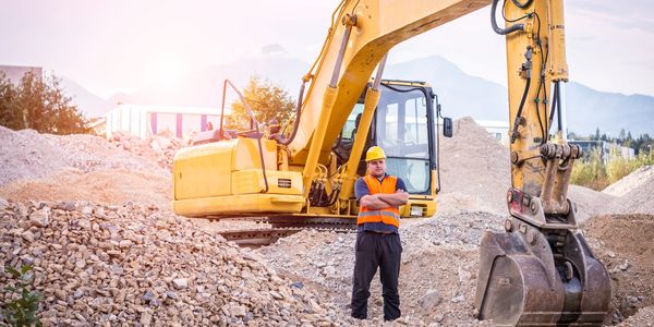 Construction worker standing with excavator amidst gravel piles at site.