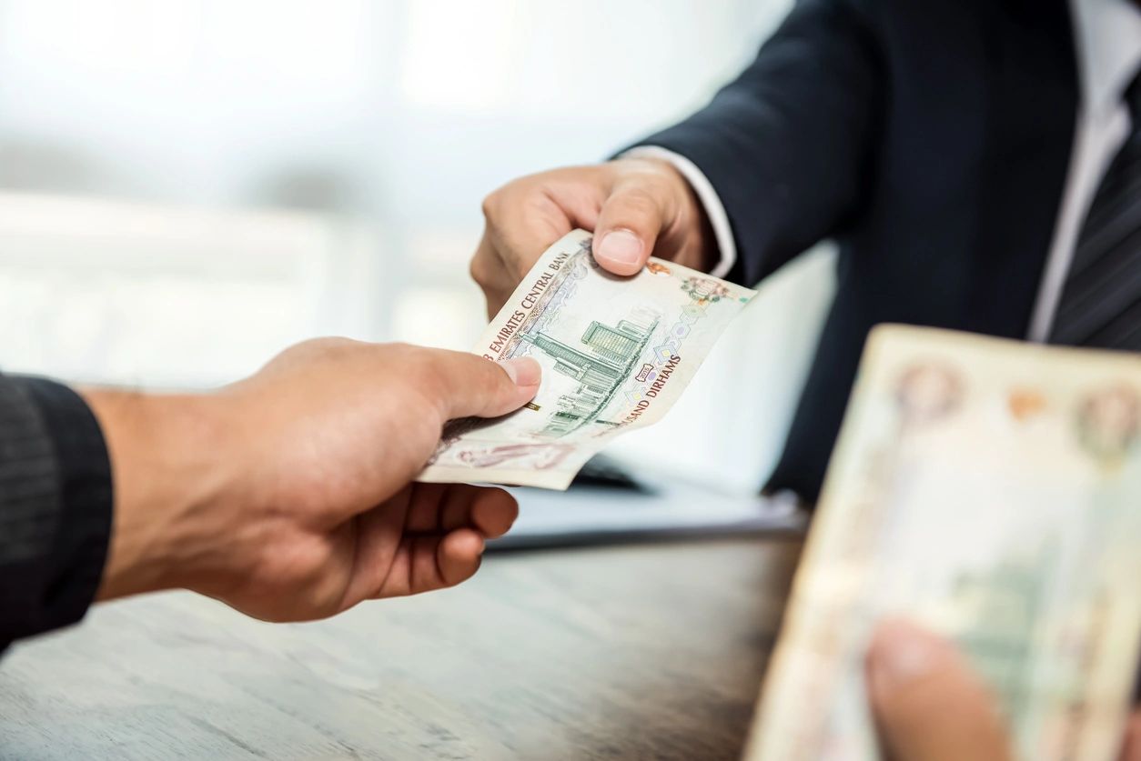 Two people exchanging UAE dirham banknotes in a business setting.