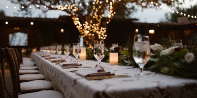 A long table setup outdoors, decorated with wine glasses, candles and fairy lights.