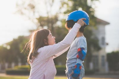 A woman helps a child put on a blue helmet and protective gear outdoors.