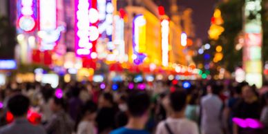 A blurred crowd walks through a brightly lit city street at night.