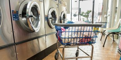 Laundry cart filled with clothes next to washing machines in a laundromat.