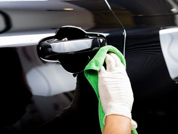 Hand in glove cleaning shiny black car door handle with green cloth.