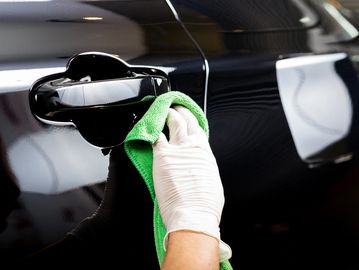 Hand in glove polishing a black car door handle with a green cloth.