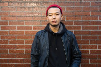 Young man in a black jacket and red beanie stands against a brick wall.
