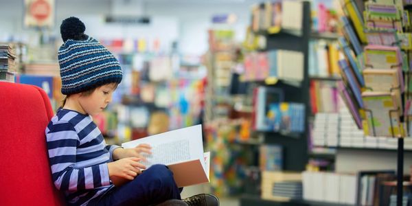  A little boy reading a book in a red chair at teh library or bookstore.